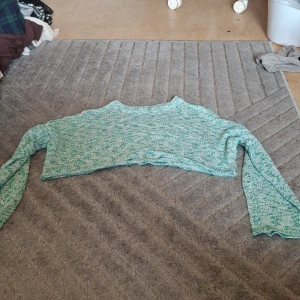 Croppad stickad tröja i mintgrönt - Superfin croppad stickad tröja i mintgrönt och vitt melerat garn. Tröjan har lång ärm och en avslappnad passform, perfekt att slänga på sig över ett linne eller klänning. Skön och trendig för dig som gillar lager på lager.