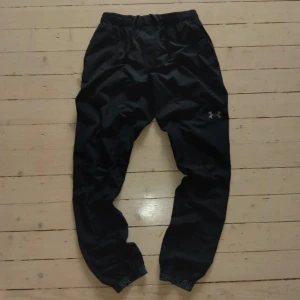 Svarta träningsbyxor från Under Armour - Svarta träningsbyxor från Under Armour med elastisk midja och snörning. Byxorna har smal passform vid anklarna och logga på låret. Perfekta för gymmet eller löprundan, tillverkade i lätt och funktionellt syntetmaterial.