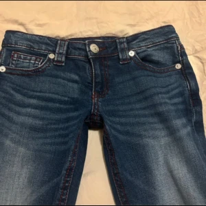 Blå bootcut jeans med röda sömmar - Snygga blå jeans med bootcut-modell och markerade röda sömmar. Jeansen har klassiska fem fickor och cool broderad detalj på bakfickorna. Låg midja och lätt slitning framtill ger en trendig look. Perfekta för dig som gillar en avslappnad men ändå stilsäker vibe.