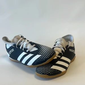 Adidas Predator svartvita fotbollsskor - Adidas Predator fotbollsskor i svart och vitt med klassiska ränder och mönstrad ovandel. Skorna har snörning och en platt gummisula som ger bra grepp på inomhusplan. Snygg och sportig design med tydlig Predator-logga på sidan.