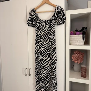 Zebramönstrad långklänning med slits - Snygg långklänning med zebramönster i svart och vitt. Klänningen har puffiga korta ärmar, fyrkantig rygg och en djup slits framtill. Figurnära passform som framhäver siluetten och ger en trendig look.