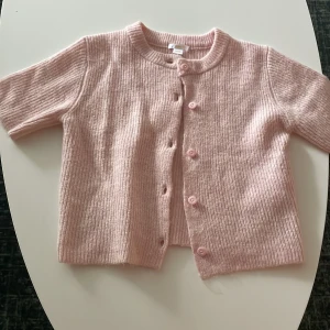 Ljusrosa ribbad kofta med knappar - Superfin ljusrosa kofta med ribbad struktur och korta ärmar. Koftan har rund halsringning och stängs med matchande knappar framtill. Perfekt att slänga över en topp för en söt och avslappnad look.