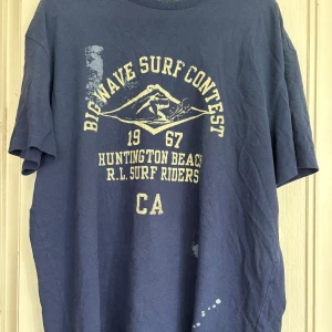 Blå t-shirt Polo Ralph Lauren M - Blå t-shirt från Polo Ralph Lauren med tryck 'Big Wave Surf Contest Huntington Beach 1967' på bröstet. Klassisk passform, kortärmad och tillverkad i mjuk bomull. Snygga vita tryckdetaljer både fram och bak för en avslappnad surfstil.