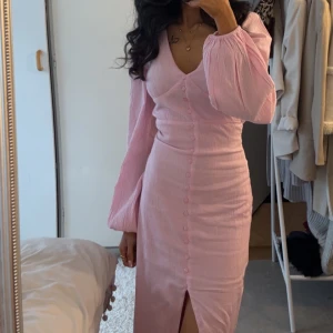 Ljusrosa långklänning med v-ringning - Nelly - Superfin ljusrosa långklänning med v-ringad hals, långa puffärmar och knappar framtill. Klänningen har en slits framtill och är figurnära i modellen. Perfekt för dig som vill ha en romantisk och trendig look. :