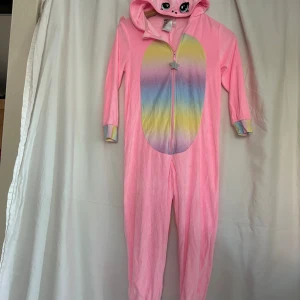 Rosa enhörningsonesie från H&M med vingar - Supergullig rosa onesie från H&M med huva som har broderade ögon, öron och regnbågsman. Framsidan har regnbågsfärgat fält och dragkedja med stjärnformad dragare. Baksidan har glittriga vingar och fluffig löstagbar svans. Be a unicorn 🦄 