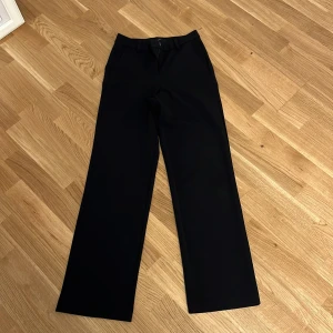 Svarta kostymbyxor med raka ben - Snygga svarta kostymbyxor med klassisk rak passform och hög midja. Byxorna har bälteshällor och sidfickor, perfekta för en clean och stilren look. Materialet känns mjukt och följsamt, vilket gör dem bekväma hela dagen.