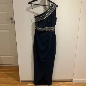 Marinblå långklänning med paljetter - Snygg marinblå långklänning med en axel och glittriga silverpaljetter upptill. Klänningen har oneshoulder-design och draperad kjol som ger en elegant look. Perfekt för bal eller festkvällar när du vill sticka ut.
