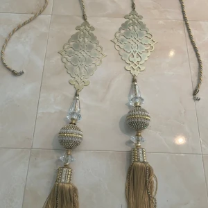 Lyxigt guldigt tassel med tofsar och kristaller - Säljer ett riktigt maffigt tassel i guld med dekorativa detaljer, gnistrande kristaller och stora tofsar. Tassel har en flätad snodd, metallplatta med orientaliskt mönster och glittrande pärlor. 