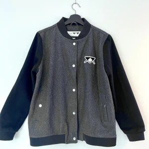 Grå bomberjacka från Junkyard L - Snygg grå och svart bomberjacka från Junkyard i storlek L. Jackan har klassisk college-look med svarta ärmar, ribbade muddar och tryckknappar framtill. Runs a bit small in size. Perfekt för dig som gillar streetstyle.