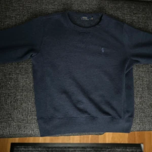 Mörkblå sweatshirt från Polo Ralph Lauren - Klassisk mörkblå sweatshirt från Polo Ralph Lauren med rund halsringning och diskret broderad logga på bröstet. Tröjan har långa ärmar och ribbade muddar vid ärmslut och nederkant. Perfekt för en chill och stilren look.