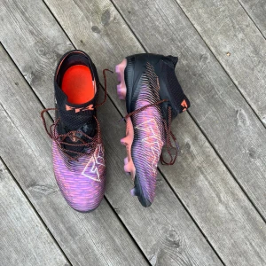 Puma fotbollsskor i svart och lila - Säljer ett par grymma Puma fotbollsskor med snyggt mönster i svart, lila och orange. Skorna har snörning, högre strumpliknande skaft och coola detaljer på sulan. Perfekta för dig som vill sticka ut på planen och ha bra grepp när du spelar. Dom är använda ett handfull gånger kanske 2-3 tre gånger stl 44 ag