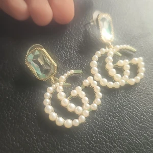  örhängen med pärlor och kristall - Statementörhängen med GG-logga i guldton, dekorerade med vita pärlor och en stor rektangulär grönskimrande kristall upptill. Lyxig och trendig accessoar som verkligen drar blickarna till sig.