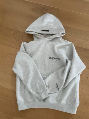Grå Essentials Fear of God hoodie - Snygg ljusgrå hoodie från Essentials Fear of God med svart logga tryckt på bröstet och ärmen. Klassisk passform med huva och ribbade muddar. Tillverkad i mjuk bomullsblandning som känns skön mot huden. Perfekt för en avslappnad och trendig look.