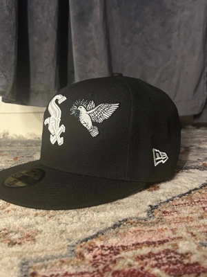 Svart New Era keps White Sox - Svart keps från New Era med broderad White Sox-logga och en vit fågel på framsidan. Klassisk flat peak och broderad New Era-logga på sidan. Snygg streetstil och passar perfekt till vardagslooken.