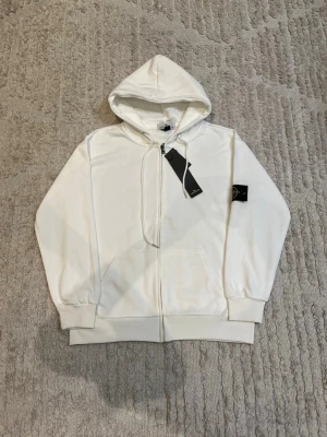 Vit zip hoodie med huva - Snygg vit hoodie med dragkedja framtill och stor huva. Tröjan har ribbade muddar vid ärmslut och nederkant samt fickor på magen. Perfekt för en clean och avslappnad stil. Materialet ser ut att vara mjukt och bekvämt, troligen bomullsblandning.