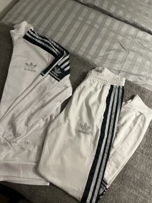 Vitt Adidas Originals träningsset - Säljer ett super snyggt vitt Adidas Originals träningsset med klassiska svarta och grå/ silvriga ränder längs ärmar och ben. Setet består av en zip-jacka och matchande byxor, båda med Adidas logga och Trefoil på bröst och lår. Materialet är mjukt och perfekt för sport eller chill.