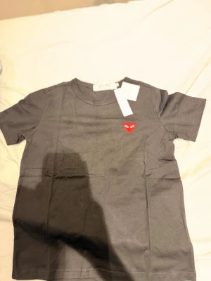 Svart t-shirt från Comme des Garçons - Svart t-shirt från Comme des Garçons Play med det ikoniska röda hjärtat broderat på bröstet. Klassisk rund halsringning och korta ärmar. Perfekt för dig som gillar stilrena plagg med en twist.