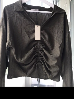 Olivgrön topp med rynk från NAKD - Trendig olivgrön långärmad topp från NA-KD med krage och snygg rynkdetalj framtill som kan justeras med snörning. Tunt, strukturerat material som ger en cool look. Perfekt att styla med jeans eller kjol.