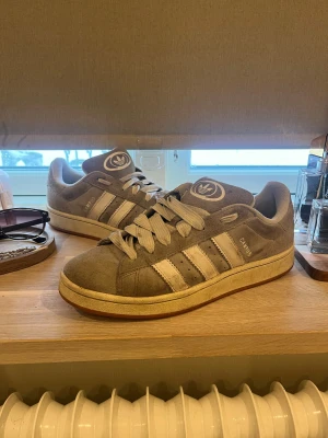 Adidas Campus grå sneakers - Snygga Adidas Campus sneakers i grå mocka med klassiska vita ränder på sidorna och gummisula. Låg modell med breda snören och Adidas-logga på plösen. Perfekta för en avslappnad streetwear-look.