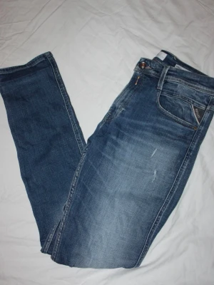 Blå Replay jeans med slitningar - Säljer ett par blå jeans från Replay med snygga slitningar och tvättad look. Klassisk femficksmodell med raka ben och detaljerade sömmar. Jeansen har en brun patch med Replay-logga bak i midjan och är tillverkade i bomull.