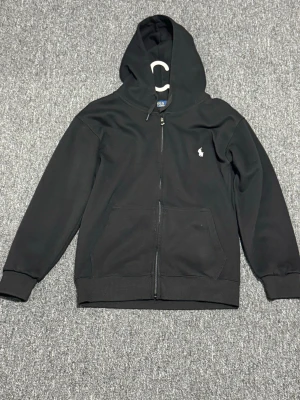 Svart hoodie från Polo Ralph Lauren - Svart hoodie från Polo Ralph Lauren med dragkedja framtill och klassisk huva. Broderad vit logga på bröstet och två fickor framtill. Tillverkad i mjukt material som är skönt att bära. Perfekt för en avslappnad och stilren look.