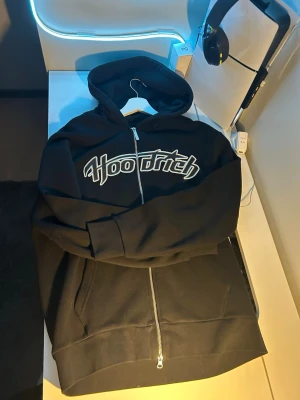 Svart Hoodrich zip hoodie - Svart hoodie med dragkedja från Hoodrich. Stor broderad logga i vitt och grått på bröstet, huva och stora fickor framtill. Perfekt för en streetwear-look och riktigt skön att bära. Materialet är mjukt och känns slitstarkt.