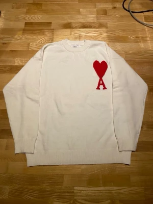 Vit stickad tröja från Ami Paris - Vit stickad tröja från Ami Paris med röd hjärtlogga och bokstaven A på bröstet. Tröjan har rund halsringning, långa ärmar och ribbstickade muddar. Perfekt för en stilren och trendig look.