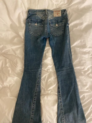 Blå bootcut jeans från True Religion - Säljer ett par blå bootcut jeans från True Religion med vita kontrastsömmar och broderade detaljer på bakfickorna. Jeansen har låg midja och klassisk femficksdesign. Materialet är denim i bomull med lätt slitning för en cool look.