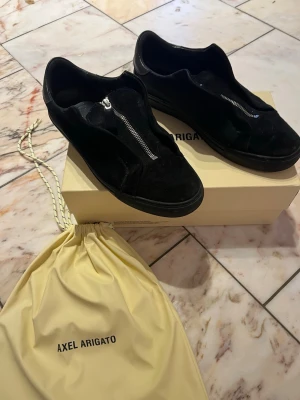 Svarta sneakers från Axel Arigato - Säljer ett par svarta sneakers från Axel Arigato med snygg dragkedja på ovansidan. Skorna har en rund tå och platt sula, tillverkade i mocka med detaljer i skinn. Kommer med original dustbag och låda. Perfekt för dig som gillar stilrena och moderna skor. Har även en söm som gått up som man ser på sista bilden.