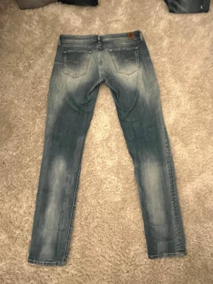 Slim fit ljusblå jeans - Säljer ett par slim fit jeans i ljusblå denim med snygga slitningar och klassiska fem fickor. Jeansen har en modern passform och är perfekta för en avslappnad stil. Materialet är bomullsdenim och de har en läderpatch bak i midjan.
