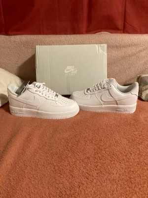 Nike Air Force 1 vita sneakers - Klassiska Nike Air Force 1 sneakers i helvitt läder med vit sula och vita skosnören. Ikonisk design med perforerad tåbox och Nike Swoosh på sidan. Perfekta för dig som gillar stilrena och tidlösa sneakers.