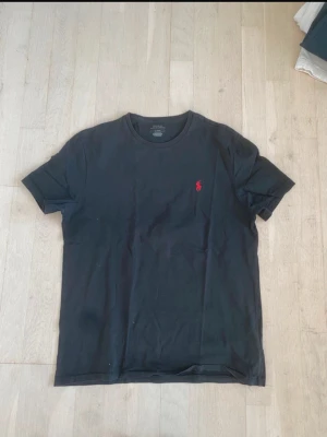 Svart t-shirt från Polo Ralph Lauren - Klassisk svart t-shirt från Polo Ralph Lauren med röd broderad logga på bröstet. T-shirten har rund halsringning och korta ärmar. Tillverkad i mjuk bomull för en skön och avslappnad stil.
