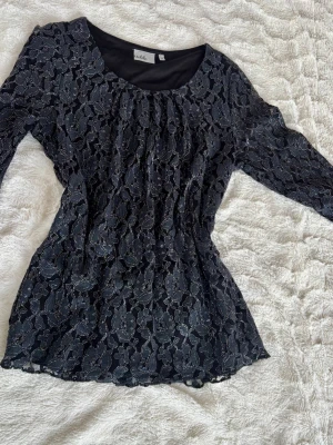 Svart spetsblus med lång ärm - Snygg svart blus från Vila med lång ärm och genombruten spets över hela toppen. Blusen har rund halsringning och ett feminint blommigt spetsmönster som ger en elegant vibe. Perfekt att styla med jeans eller kjol för en trendig look.