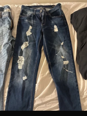 Mörkblå slitna jeans med hål - Säljer ett par mörkblå jeans med tydliga slitningar och hål på både fram- och baksida. Jeansen har klassisk femficksmodell och raka ben. Perfekta för en avslappnad och trendig look.