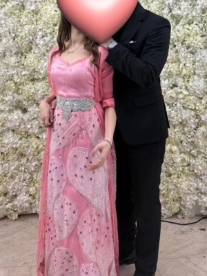 Rosa kurdisk lawandi med paljettdetaljer - Elegant rosa långklänning med glittriga paljettdetaljer och ett brett, dekorerat bälte i midjan. Klänningen har v-ringad hals, halvtransparent tyg med mönster. Perfekt för speciella tillfällen. Klänningen har används i några timmar och passar storlek 34-40. Pris kan diskuteras vid snabb affär.