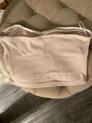 Beige ribbad bandeau tubtopp - Säljer en beige ribbad bandeau tubtopp med twistad detalj framtill. Toppen är ärmlös och har en croppad passform, perfekt för varma dagar eller som layering piece. Materialet är mjukt och toppen har resor band.