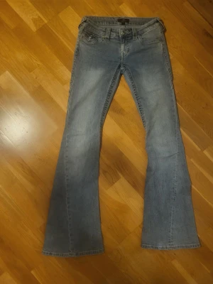 Blå bootcut jeans från True Religion  - Säljer ett par ljusblå bootcut jeans från True Religion med klassisk femficksdesign och snygga detaljer på bakfickorna. Jeansen har en något utsvängd passform nedtill och är tillverkade i ett mjukt denimtyg. Perfekta för dig som gillar retrostil och vill ha ett par jeans med cool attityd.