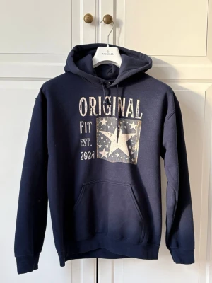 Mörkblå hoodie med tryck och huva - Mörkblå hoodie från SCANDI med stor huva och magficka. Framsidan har ett coolt tryck med texten 'ORIGINAL FIT EST. 2024' och en stjärna i en ruta fylld med mindre stjärnor. Tillverkad i mjukt material, perfekt för chill dagar.