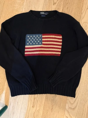 Ralph Lauren stickad - Snygg mörkblå stickad tröja från Polo Ralph Lauren i 100% bomull. Tröjan har ett stort amerikanskt flaggmotiv på bröstet och ribbade muddar vid ärmslut och nederkant. Perfekt för dig som gillar klassisk preppy stil.
