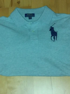 Grå piké från Polo Ralph Lauren - Klassisk grå pikétröja från Polo Ralph Lauren med stor mörkblå logga broderad på bröstet och siffran 3 på ärmen. Tröjan är långärmad, krage och knappar framtill. Perfekt för en clean och sportig stil. Tröjan är I storlek 14-16 alltså 160. Hör av er vid intresse eller om ni har några frågor!