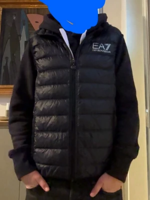 Svart dunväst från EA7 Emporio Armani - Snygg svart dunväst från EA7 Emporio Armani med quiltad design och dragkedja framtill. Västen har en diskret logga på bröstet och glänsande yta. Perfekt att slänga över en hoodie för en cool och sportig look.