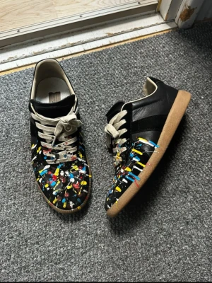 Maison margiela gats  - Unika svarta sneakers från Maison Margiela med färgstänk i blått, gult, vitt och rött. Skorna har beige snören, klassisk låg siluett och gummisula. Tillverkade i läder med en konstnärlig vibe som sticker ut.