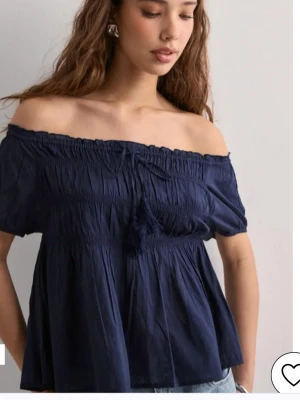 Mörkblå offshoulder blus - Snygg mörkblå blus med offshoulder-modell och korta ärmar. Toppen har smock upptill och en dekorativ knytning framtill. Perfekt för en trendig och avslappnad look.