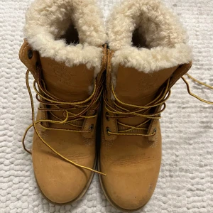 Timberland boots med fårskinnsfoder - Klassiska Timberland boots i beige nubuck med grov sula och snörning i gult och brunt. Insidan är fodrad med mjukt, vitt fårskinn som håller dig varm. Ikonisk Timberland-logga på sidan och robusta metalldetaljer vid snörningen. Perfekta för kalla dagar.