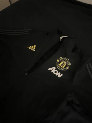 Manchester United adidas träningsjacka - Säljer en svart träningsjacka från adidas med Manchester Uniteds klubbmärke på bröstet och AON-logga. Jackan har dragkedja, adidas-logga i gult och klassiska ränder på ärmarna. Perfekt för dig som älskar fotboll och vill ha en snygg sportig look. Knappt använd 