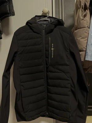 Svart vindjacka Sail Racing - Svart vindjacka från Sail Racing med quiltade partier och huva. Jackan har dragkedja framtill, två sidofickor och logga på bröstet samt detaljer på ärmen och huvan. Materialet är lätt och vindtätt, perfekt för blåsiga dagar.