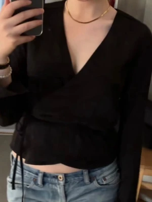 Svart omlottopp - Snygg svart omlottopp med långa ärmar och knytband i sidan. Toppen har en djup v-ringning och sitter croppad i midjan. Perfekt att styla med jeans eller kjol för en trendig look.