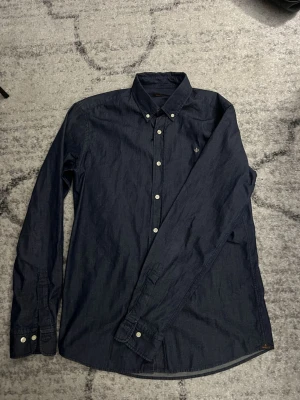 Mörkblå skjorta från Morris - Säljer en stilren mörkblå skjorta från Morris med klassisk krage och vita knappar. Skjortan har lång ärm och en liten broderad logga på bröstet. Perfekt till jeans eller chinos för en clean look.