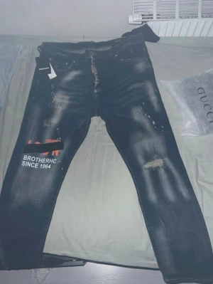 Dsquared2 blå slitna jeans - Säljer ett par blå jeans från Dsquared2 med slitningar, tryck och textdetaljer på vänster ben. Jeansen har en tvättad look, vita och röda detaljer samt klassisk femficksmodell. Materialet är denim och de har en modern, trendig passform.