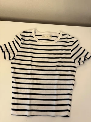 Randig ribbad t-shirt  - Vit ribbad t-shirt från Pull&Bear med svarta horisontella ränder. Perfekt för en avslappnad och trendig stil. Liten i storleken 